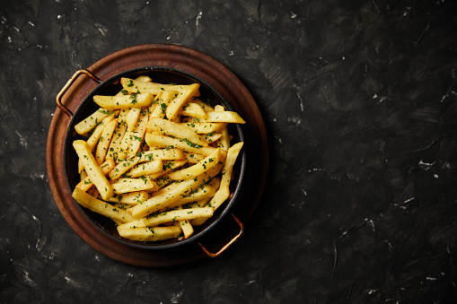 french fries seasoned with parsley and garlic served in a frying pan on a wooden plate on a dark background