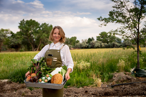 Pretty Blond Woman in Her twenties Standing outside near a field displaying a tray of organic food products such as eggs, milk and vegetables from an organic market