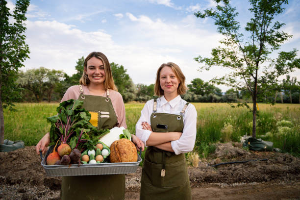 duas mulheres com uma cesta de produtos orgânicos, leite, ovos e outras compras posando orgulhosamente em frente a um campo de feno em sua fazenda sustentável de varejo orgânico - organic farmers market market vegetable - fotografias e filmes do acervo