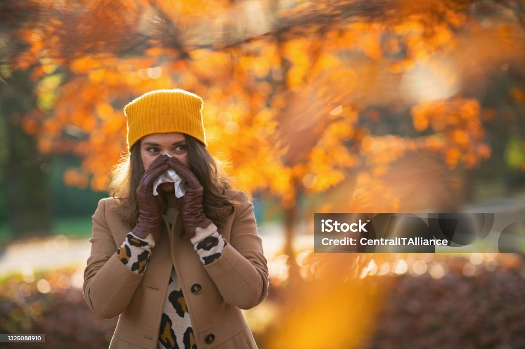 modern 40 years old woman in brown coat and yellow hat Hello autumn. modern 40 years old woman in brown coat and yellow hat with napkin blowing nose outdoors in the city park in autumn. Autumn Stock Photo