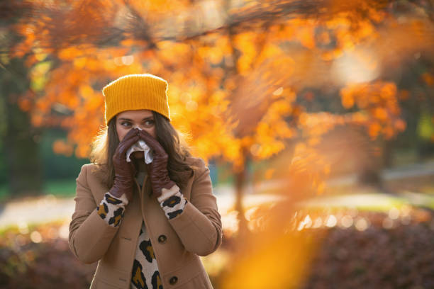 modern 40 years old woman in brown coat and yellow hat Hello autumn. modern 40 years old woman in brown coat and yellow hat with napkin blowing nose outdoors in the city park in autumn. allergy stock pictures, royalty-free photos & images