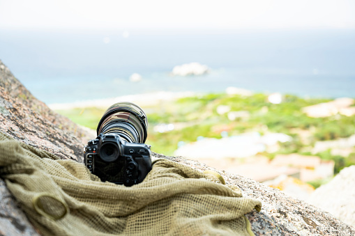 (Selective focus) Professional camera with a super telephoto lens with camouflage cover on a rocky mountains. Paparazzi equipment, Sardinia, Sardinia, Italy.