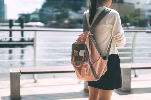 Young adult female college student backpack rear view. Rear view of young adult female college student wear sweater and pink color backpack. Standing at park area with river and city backgroud on day time. female high school student stock pictures, royalty-free photos & images