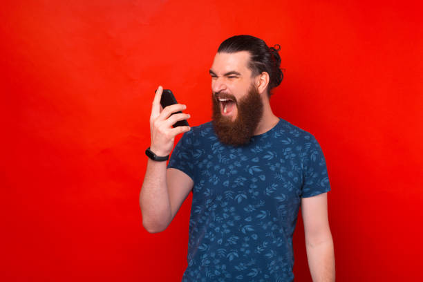 istock Young bearded hipster man screaming at smartphone over red background 1325187275