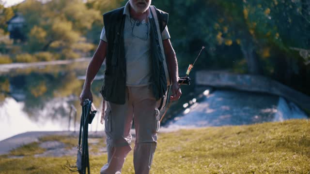 Senior 70s man walks by the river carrying a fishing rod and fishing equipment