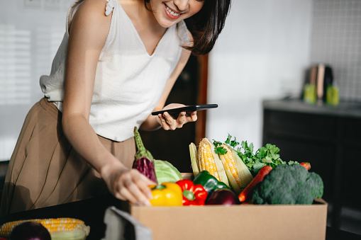 Beautiful smiling young Asian woman doing home delivery grocery shopping online with mobile app device on smartphone at home, with a box of colourful and fresh organic vegetables and fruits on the table