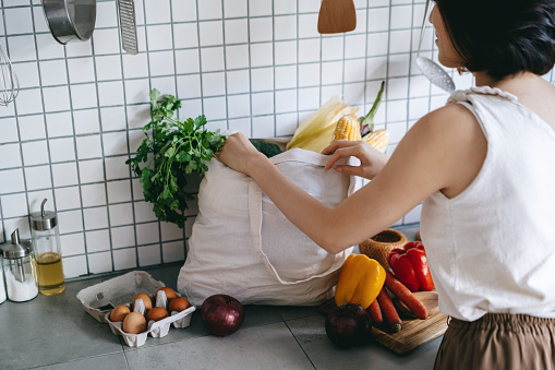 Young Asian woman coming home from grocery shopping and taking out fresh fruits and vegetables from a reusable shopping bag on the kitchen counter at home