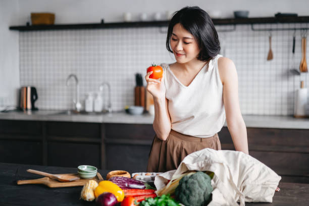 jeune femme asiatique rentrant de l’épicerie et sortant des fruits et légumes frais d’un sac réutilisable sur le comptoir de la cuisine. elle prévoit préparer un repas sain avec des produits frais - asian meal photos et images de collection