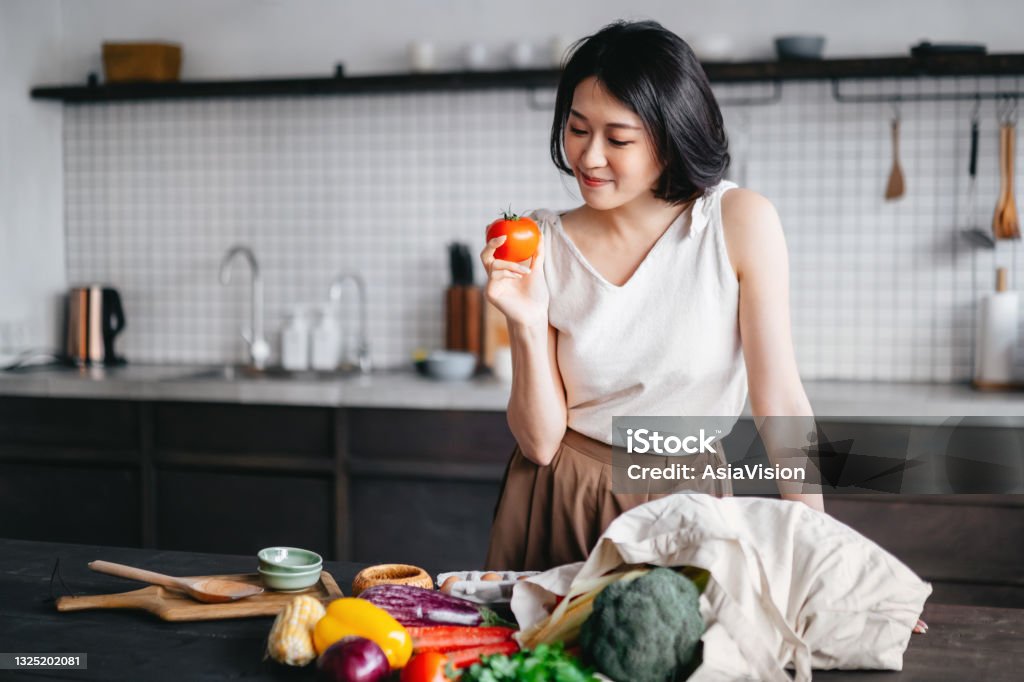 Young Asian woman coming home from grocery shopping and taking out fresh fruits and vegetables from a reusable shopping bag on the kitchen counter. She is planning to prepare a healthy meal with fresh produces Nutritionist Stock Photo