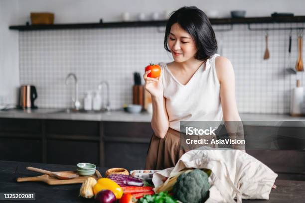Young Asian Woman Coming Home From Grocery Shopping And Taking Out Fresh Fruits And Vegetables From A Reusable Shopping Bag On The Kitchen Counter She Is Planning To Prepare A Healthy Meal With Fresh Produces Stock Photo - Download Image Now