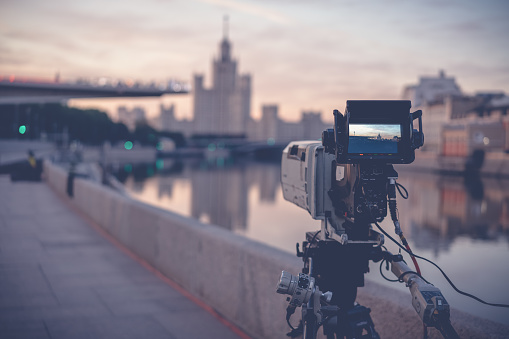 A professional video camera stands on a tripod recording the city and the river at dawn