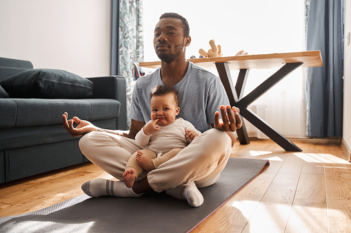 Calm multiracial man in casual clothes meditating with his son and sitting on lotus pose at home. Yoga and wellness concept. Stock photo