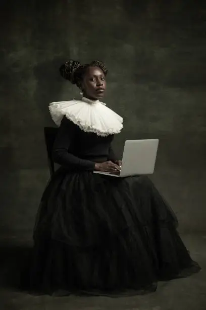 Photo of Portrait of medieval African young woman in black vintage dress with big white collar posing isolated on dark green background. Using laptop