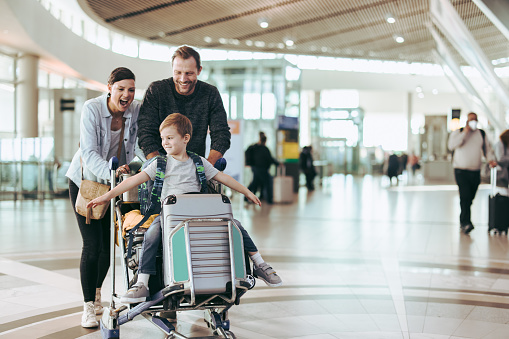 Couple happily pushing the trolley with their son at airport. Child enjoying sitting on luggage trolley while parents pushing it at airport.