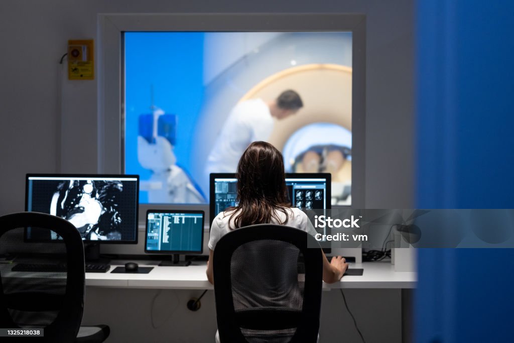 Doctor analyzing MRI results in office next to scanner Vertical rear view photo of an unrecognizable female doctor analyzing MRI results in an office. MRI scanner and doctor in the background. Diagnostic Medical Tool Stock Photo