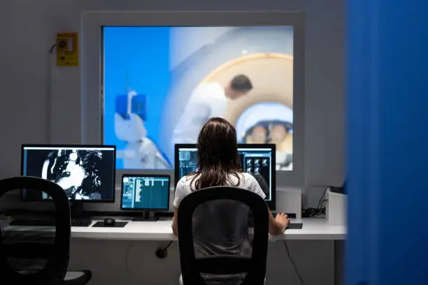 Vertical rear view photo of an unrecognizable female doctor analyzing MRI results in an office. MRI scanner and doctor in the background.