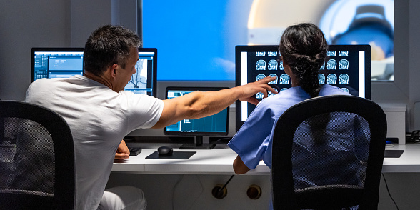 Back view of two doctors analyzing MRI scan results. Man pointing at images on monitor screen. Magnetic resonance imaging technology in specialized medical clinic.