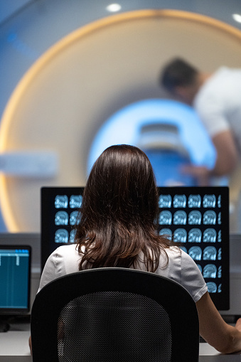 Vertical rear view photo of an unrecognizable female doctor analyzing MRI results in an office. MRI scanner and doctor in the background.