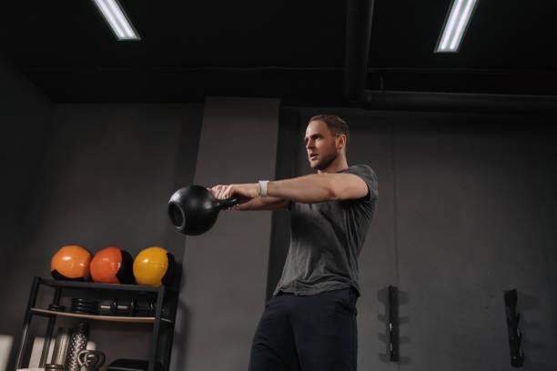 Athlete doing kettlebell exercise at gym Strong muscular athlete doing kettlebell swings at gym, outfit shot on grey gym background with equipment. Weightlifter doing workout indoors, exercising. Instructor warming up circuit training stock pictures, royalty-free photos & images