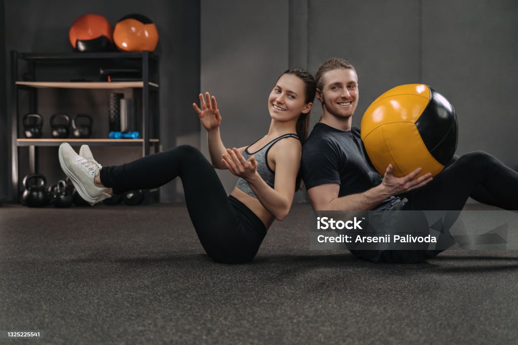 Couple exercising together with a ball Happy couple training together at dark gym with professional sport equipment on background. Fit man and sporty woman doing abs exercise together using medicine ball, passing it each other High-intensity interval training Stock Photo