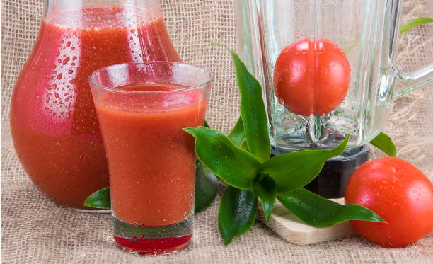Tomato juice in glass pitcher under water drops, blender, greens, two raw tomatoes on burlap background stock photo