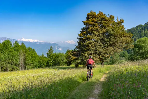 nice, active senior woman riding her electric mountain bike at the slopes of Monte Velo in the Sarca Valley with Monte Adamello in background, Garda lake mountains, Trentino, Italy