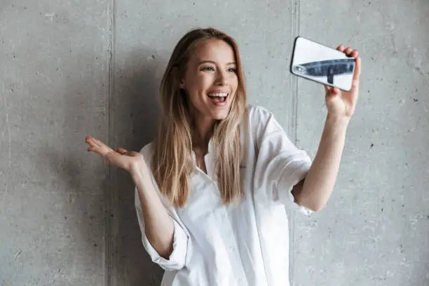 Portrait of a cheerful young girl taking a selfie while standing over gray wall background