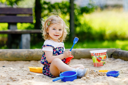 Cute toddler girl playing in sand on outdoor playground. Beautiful baby in red trousers having fun on sunny warm summer day. Child with colorful sand toys. Healthy active baby outdoors plays games.