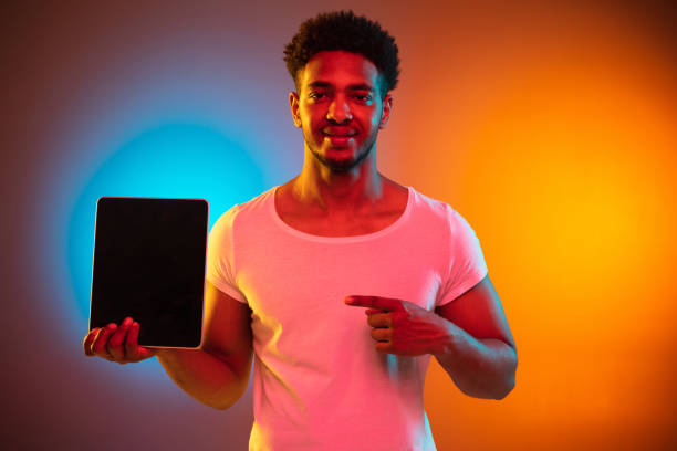 istock African-american man's portrait isolated on gradient studio background in neon light. Beautiful model with tablet. 1325244669