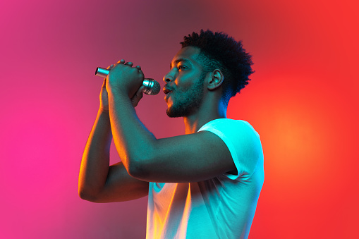 African handsome guy singing into the microphone in the studio on a neon background. Music concept. Young joyful attractive guy improvising.