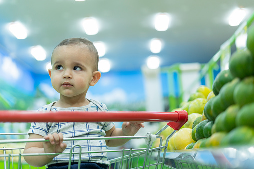 One-year-old baby accompanies her mother to do the grocery shopping riding in the grocery shopping cart while she touches the fruits