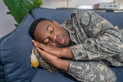 Tiredness, sleep. Young adult african american man in military uniform sleeping on sofa with hands under head at home