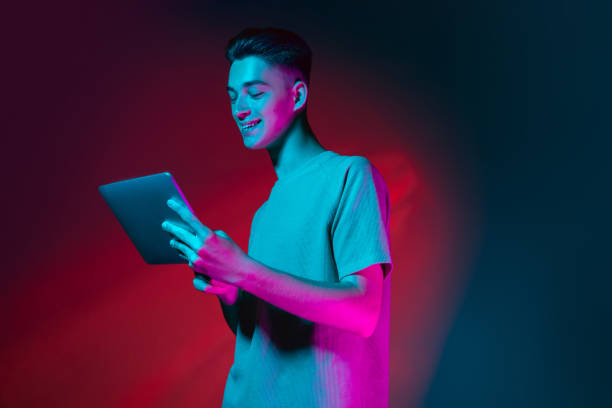 Portrait of Caucasian young man with tablet, device isolated on dark red studio background in neon pink light. Concept of human emotions, facial expression. Using tablet. Young man with gadget, device isolated on dark red background in neon pink light. Handsome male model in casual style. Concept of emotions, facial expression, youth. Copy space for ad. neon portrait boy stock pictures, royalty-free photos & images
