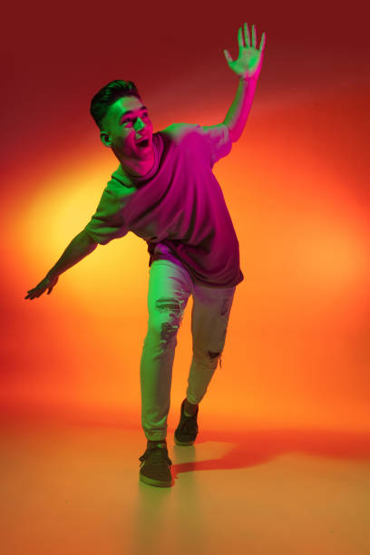 Portrait of Caucasian young man dancing, dancing isolated on orange studio background in neon light. Concept of human emotions, facial expression. Fun, joy. Excited happy young man dancing isolated on orange background in neon pink light. Handsome male model in casual style. Concept of emotions, facial expression. Copy space for ad. neon portrait boy stock pictures, royalty-free photos & images