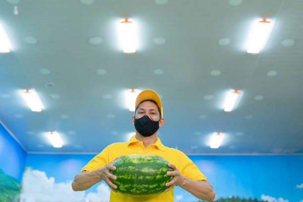The employee of the Latin greengrocer from Bogota Colombia, between 40 and 49 years old, wears his uniform and mask while looking at the camera in a portrait and smiling while arranging the fruits.