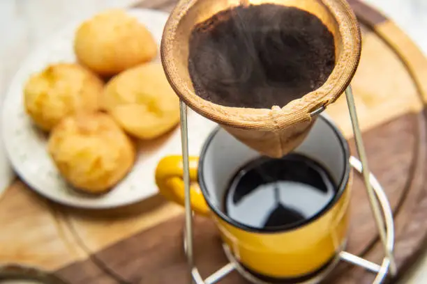Goiânia, Goias, Brazil – June 24, 2021: Straining coffee into the fabric strainer with a yellow mug underneath. Plate with cheese buns in the background. Preparing breakfast.