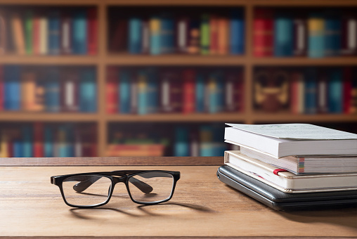Eyeglasses and stacked books with laptop on wooden desk with blurred bookshelf in home office room