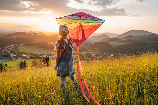 Little girl enjoying the sunset on the meadow grass and preparing colorful rainbow kite toy for flying. Happy childhood moments or outdoor time spending concept image.
