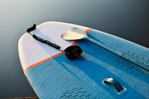Paddle board and surf board with paddle on blue water surface background close up. Surfing and SUP boarding equipment in sunset lights close-up. Paddleboard with a paddle on a wooden pier in the lights of sunset on the background of a forest lake close-up. Outdoor water sports. Surfing lifestyle backgrounds.