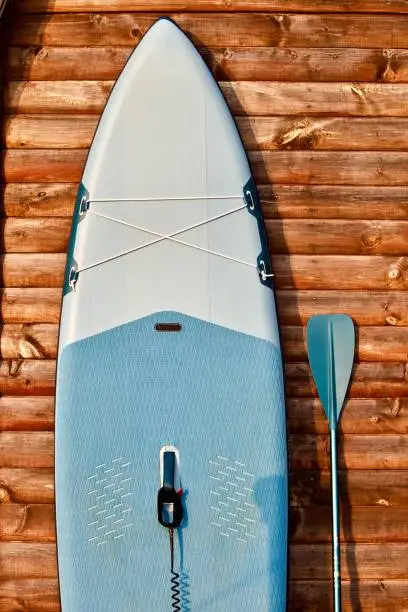 Paddle board and surf board with paddle on brown wooden wall background close up. Surfing and SUP boarding equipment in sunset lights close-up. Paddleboard with a paddle on a wooden pier in the lights of sunset on the background of a forest lake close-up. Outdoor water sports. Surfing lifestyle backgrounds.