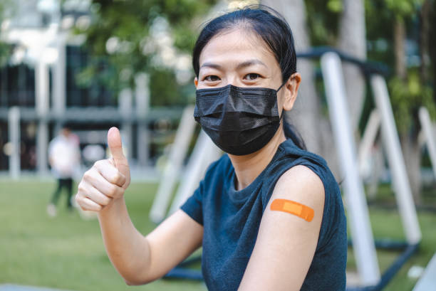 Asian woman showing thumb up after getting Covid-19 vaccine Portrait of smiling Asian woman wearing protective face mask looking at camera and showing thumb up after receiving the Covid-19 vaccine while sitting in outdoor park medical injection photos stock pictures, royalty-free photos & images