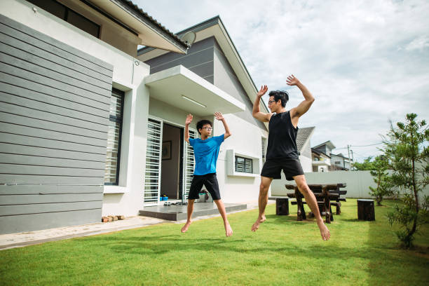 Asian chinese father doing jumping jack together with his son in front yard of house wide shot of young family exercise together every morning child jumping jack stock pictures, royalty-free photos & images