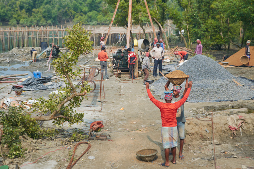 West Bengal, 01-24-2021: Manual labourers are busy at a rural construction site at Sagar Island. Few workers are seen carrying basket full of sand on their head. Heaps of sand, stone chip, iron beam and other construction material are seen at the site.