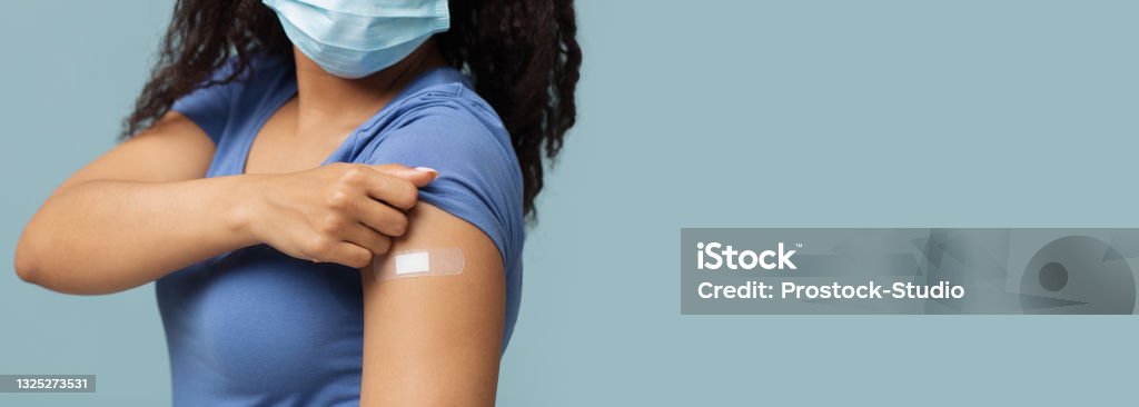 Antiviral vaccination. African american woman showing plaster on shoulder after covid-19 vaccine injection, panorama Antiviral vaccination. Unrecognizable african american woman in protective mask showing plaster on shoulder after covid-19 vaccine injection, standing over blue background, panorama with free space Vaccination Stock Photo