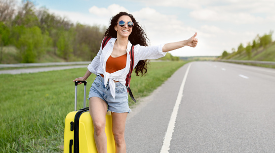 Cheerful young lady thumbing her ride, standing with suitcase on highway, trying to catch passing car, hitchhiking in countryside, panorama with copy space. Summertime adventre concept