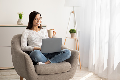 Favorite work at home, online study and self-isolation during COVID-19 outbreak. Happy relaxed young arabian lady holding laptop and cup of hot drink sitting in armchair, in living room interior