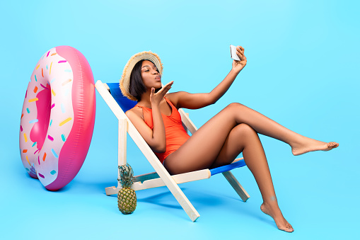 Flirty black woman chatting with her boyfriend on smartphone, blowing air kiss at webcam, making selfie while sitting in lounge chair on beach, blue studio background. Full length portrait