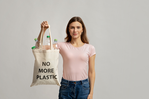 Young Smiling Female Carrying Eco Tote Bag With No More Plastic Incription Filled With Empty Bottles For Recycling, Happy Millennial Woman Enjoying Zero Waste Living, Standing On Grey Background