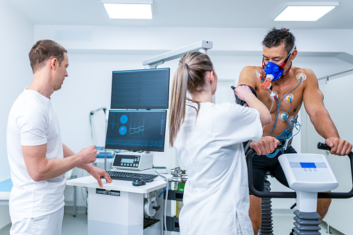 Doctor adjusting blood pressure strap on an athlete's arm during biometric testing on an exercise bike in a lab.