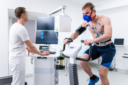 Doctor using a computer to monitor an athlete riding an exercise bike during biometric testing in a lab.